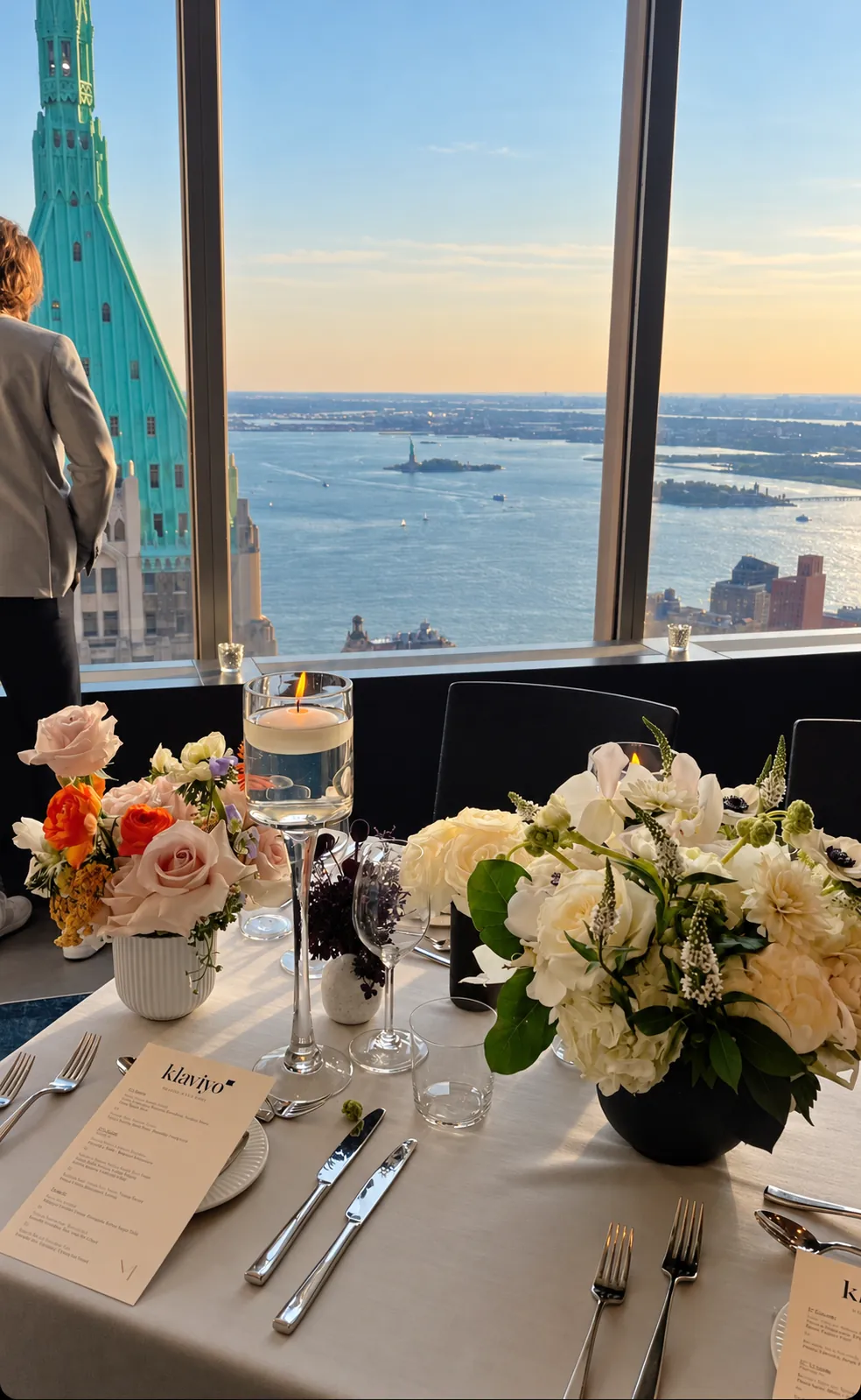 An executive dinner table with florals and candles beside floor-to-ceiling windows, sixty floors above New York Harbor at sunset