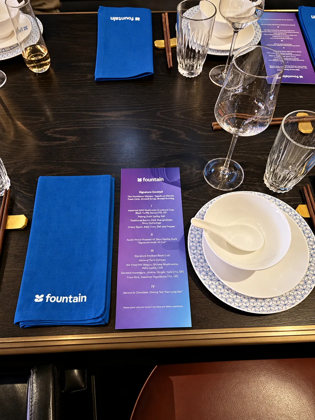 A dark lacquered dinner table set with blue linen napkins, a printed menu, porcelain and crystal glassware