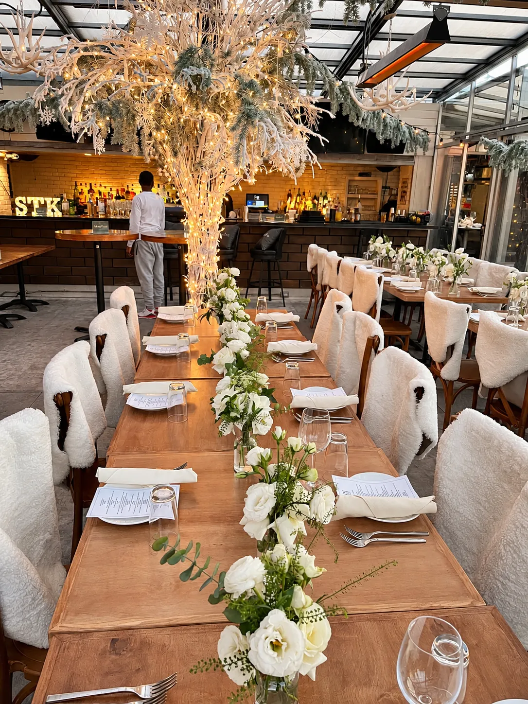A long candlelit dinner table beneath white-flocked trees, chairs draped in sheepskin, set for ten guests